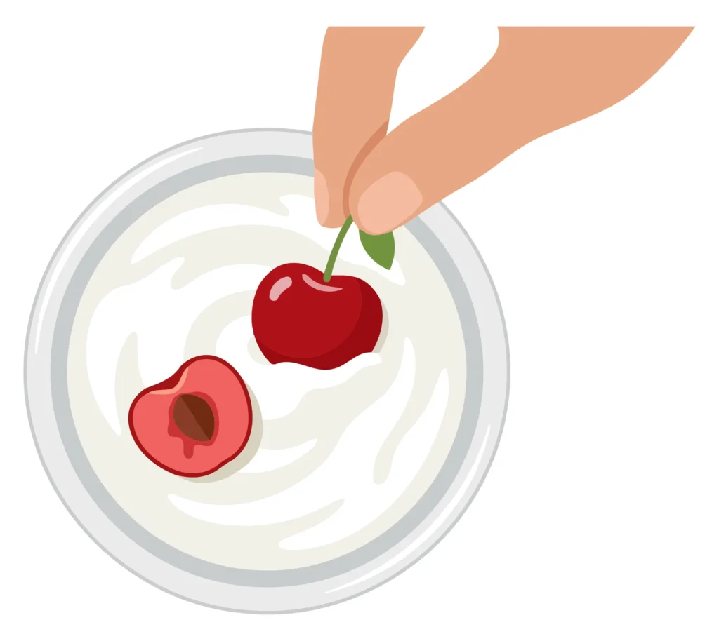 A hand places a whole red cherry into a bowl of yogurt with a light background.